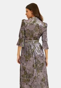 Largo vestido de terciopelo en gris con un patrón floral verde. Presenta mangas abullonadas y una cintura ajustada, que se extiende hasta una falda plisada.