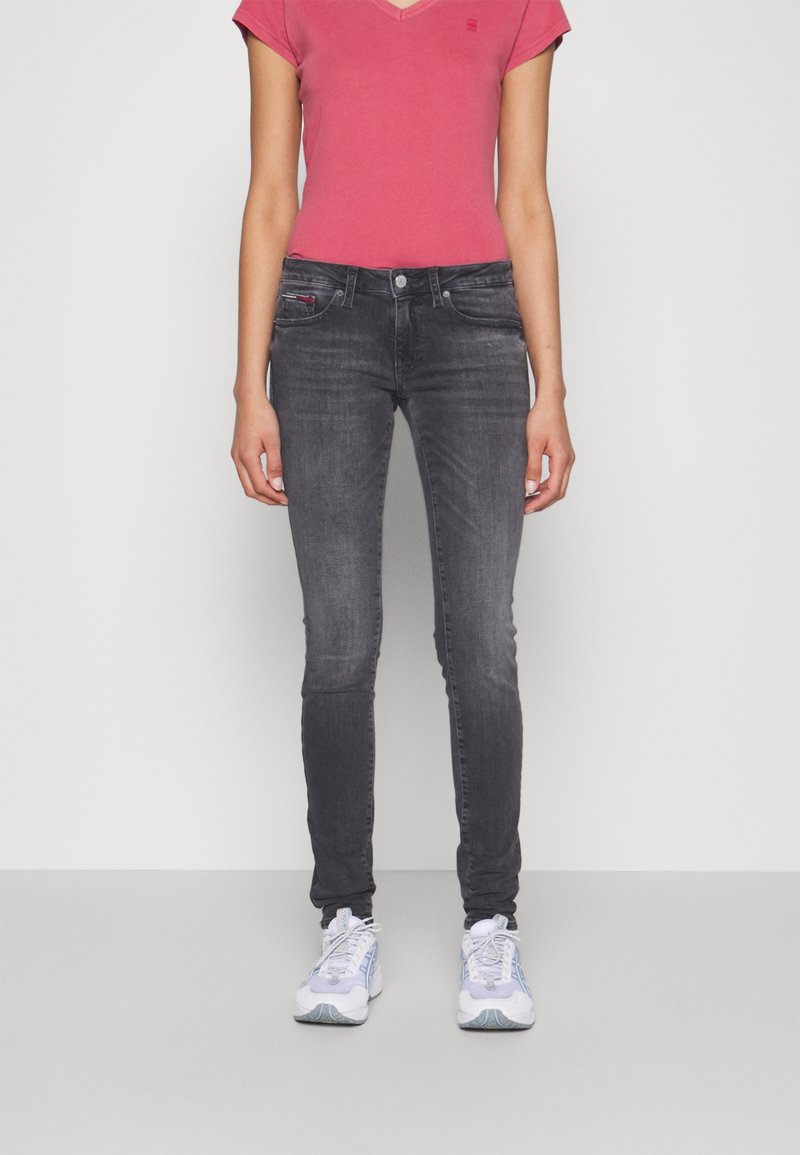 Jeans ajustados en denim gris oscuro, combinados con una camiseta de cuello en V rosa ajustada. El modelo lleva zapatillas blanca y azul claro. Diseño simple y casual.