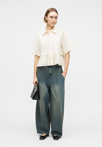 Γυναίκα που φοράει λευκό μπλουζάκι με κοντά μανίκια και πλισέ τελείωμα, φαρδιά ξεβαμμένα μπλε τζιν παντελόνια, μαύρα μοκασίνια, κρατώντας μια μικρή μαύρη τσάντα.