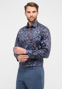 Mann passt den Bund seines dunkel gemusterten Paisley-Hemdes an, das er mit blauen Hosen kombiniert, vor einem schlichten hellen Hintergrund.
