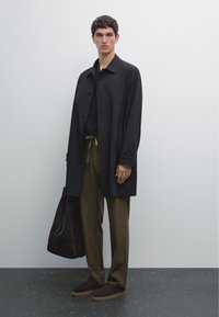Manteau noir à boutons sur un haut sombre, pantalon vert olive et chaussures en daim marron. Portant un sac fourre-tout marron foncé, debout sur un sol texturé.