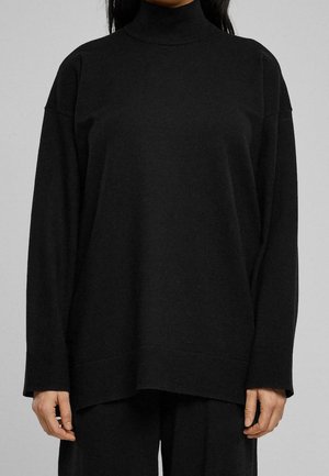 Kobieta ubrana w luźny czarny sweter z golfem i długimi rękawami oraz dopasowane czarne spodnie na jednolitym tle.