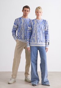 Pulls en tricot surdimensionnés avec des motifs bleu et blanc, associés à des pantalons beiges et bleu clair. Tissu texturé et design décontracté.