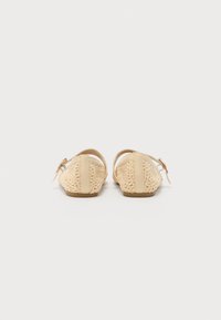 Zapatos planos tejidos en color beige con un diseño de espalda abierta, que cuentan con una correa de cuero suave y una pequeña hebilla decorativa.