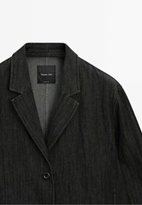 Veste noire tissée avec col à crans, fermeture à un seul bouton et tissu texturé. Étiquette visible à l'intérieur. Design minimaliste, coupe ajustée.