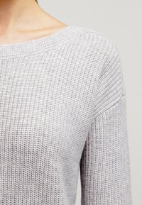 Světle šedý pletený svetr s širokým výstřihem, texturovaným vzorem a spadlými ramenními švy, vyrobený z měkkého, prodyšného materiálu.