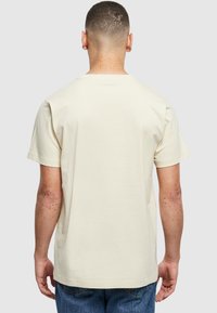 Camiseta de algodón color crema, de mangas cortas, con cuello redondo, corte relajado, textura suave, costuras visibles a lo largo del dobladillo y las mangas.