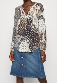Blusa a maniche lunghe in blu navy e crema, con intricati motivi paisley e floreali. Abbinata a una gonna di jeans con bottoni dorati.