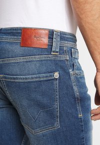 Mörkblå denimjeans med en brun läderapplikation på midjan. Har två bakfickor med gul sömnad och metallbeslag.