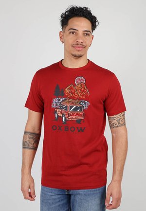 T-shirt rouge en coton avec un design graphique de montagnes et d'un véhicule. Col rond et manches courtes. Accentué par une illustration d'arbre.