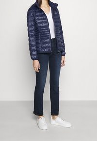 Chaqueta acolchada navy con diseño de acolchado vertical, capucha y cierre de cremallera, combinada con jeans oscuros y zapatillas blancas, de pie frente a un fondo neutro.