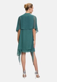 Abito di chiffon turchese con un design a strati, maniche a sbuffo corte e orlo fluente. Abbinato a sandali con tacco alto argentati.