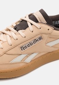 Beige leather sneaker with white side stripes, textured dark brown accents, and a rubber sole. Features Reebok logo on the side.