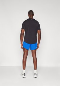 Fekete sportpóló, kék rövidnadrág és fehér sneakers. Sportos öltözet rövid ujjakkal, kerek nyakkal, és a rövidnadrágon látható logóval.