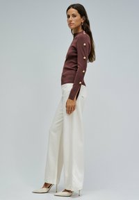 Suéter de punto marrón oscuro con acentos de botones dorados en las mangas, combinado con pantalones de pierna ancha blancos y tacones beiges con punta.