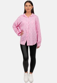 Camicia button-up rosa chiaro con motivi a cuore bianchi, maniche lunghe e colletto, abbinata a leggings neri e sneakers bianche.