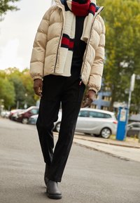 Persona caminando por la calle de la ciudad con una chaqueta acolchada beige, pantalones negros, botas negras y una bufanda color crema con rayas rojas y marinas.