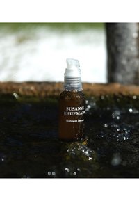 Brązowa szklana butelka z dozownikiem przystosowanym do pompowania, oznaczona "Serum odżywcze Susanne Kaufmann". Otoczona wodą z bąbelkami, odbijającą światło.