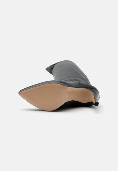 Botte noire en cuir à talon aiguille montant au genou, posée sur le côté, montrant une semelle beige lisse et un bout pointu sur fond blanc.