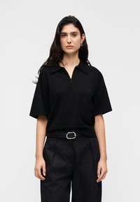 Μαύρο πόλο μπλουζάκι με κοντό μανίκι, κλείσιμο με μισή φερμουάρ και γιακά, συνδυασμένο με μαύρο παντελόνι ψηλόμεσο και ζώνη με ασημένια αγκράφα.