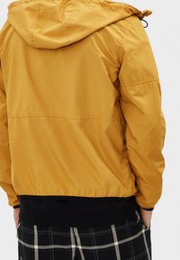 Žlutá bunda s kapucí, elastickým lemem, lehkým materiálem a hladkou texturou. Nositelná přes černobílé kárované kalhoty.