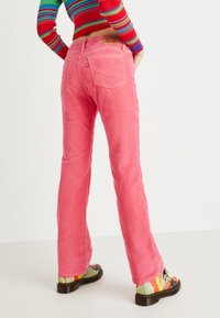 Jeans acampanados rosas con cintura alta y bolsillos traseros, hechos de mezclilla. Combinados con botas estampadas que presentan colores brillantes y una suela gruesa.