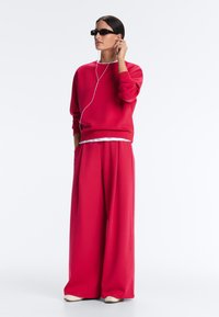 Femme portant un pantalon large rouge et un sweat-shirt, lunettes de soleil et écouteurs, debout avec une main dans la poche.