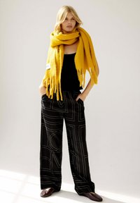 Übergroßer gelber Schal, schwarzes Tanktop, schwarze weite Hose mit weißen geometrischen Mustern und dunkelbraunen Schuhen. Weiche Texturen und kräftige Farben.