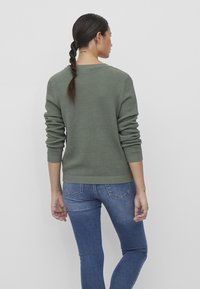 Pull en tricot vert avec un motif texturé, col rond et ourlets côtelés, associé à un jean bleu ajusté. Coiffure à tresses visible.