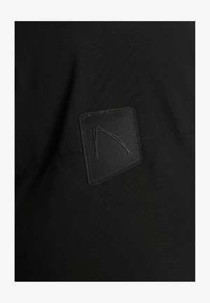 Czarna tkanina o gładkiej fakturze z kwadratową, wtkowaną metką. Metka ma wytłaczany wzór stylizowanych łuków w ciemnym odcieniu.