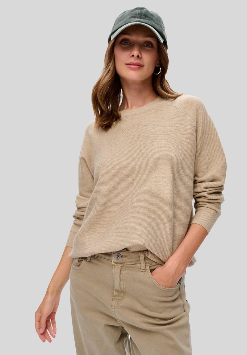 Beige geribbelde trui met lange mouwen, gecombineerd met lichtbeige broek. Het model draagt een groene pet. De achtergrond is grijs.