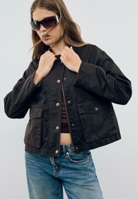 Veste noire cropped avec deux poches avant, fermeture �à pression et à zip, associée à un jean bleu. Le tissu est lisse, avec une finition mate.