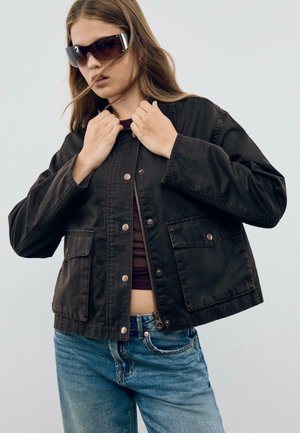 Zwarte cropped jack met twee voorzakken, drukknoop- en ritsclosure, gecombineerd met blauwe jeans. De stof is glad, met een matte afwerking.