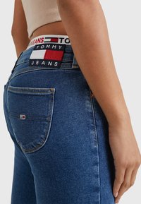 Niebieskie dżinsy z wysokim stanem, z markowym paskiem w kolorze czerwonym, białym i granatowym. Zawierają tylne kieszenie i widoczne detale szwów.