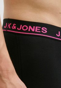 Schwarze Boxer Shorts mit einem pinken elastischen Bund, auf dem "JK & JONES" in fetten Buchstaben gedruckt ist; die glatte Stoffstruktur ist sichtbar.