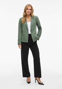 Blazer verde de tela suave, cierre de un solo botón, dos bolsillos frontales. Combinado con pantalones negros ajustados y tacones negros con acentos dorados.