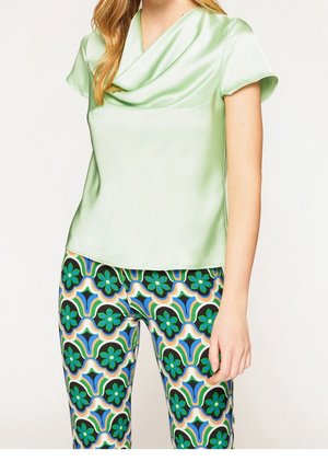 Vrouw die een lichtgroene satijnen blouse met korte mouwen en nauwsluitende broek draagt met een blauw, groen en zwart bloemengeometrisch patroon.