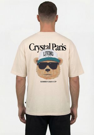 Man draagt een beige T-shirt met een beer die een zonnebril en een pet draagt, tekst "Crystal Paris" boven en "Summer 2026 © CP" onder de beer.