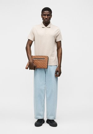 Hombre con polo beige de manga corta, vaqueros anchos de pierna ligera azul claro, zapatos negros, sosteniendo un bolso de mano marrón de cuero con correa en la muñeca, de pie sobre fondo blanco.