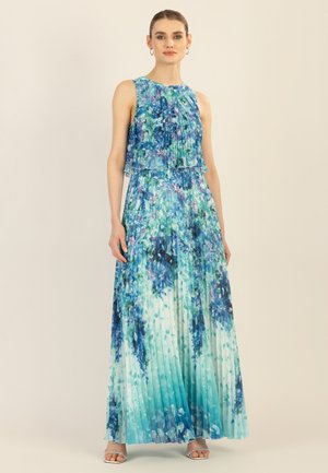 Frau trägt ein ärmelloses, plissiertes Maxikleid mit blau-grünen Blumenmustern, stehend vor einem hellbeigen Hintergrund.