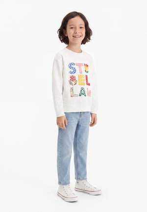 Enfant aux cheveux foncés souriant, portant un sweat blanc avec des lettres fleurs colorées et un jean bleu clair avec des baskets montantes blanches.