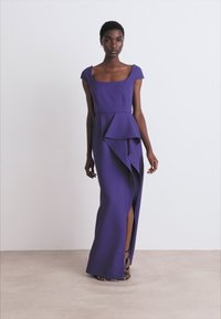 Langes lila Kleid mit kurzen Ärmeln, strukturiertem Oberteil und großem Vorderfall. Ein seitlicher Schlitz zeigt die Beine und schwarze Riemchenschuhe.