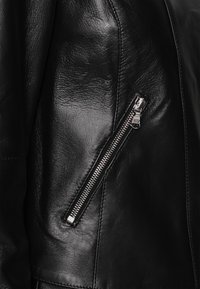 Giubbotto in pelle nera con cerniera metallica argentata, texture liscia, design angolare e cuciture di rifinitura sui lati.