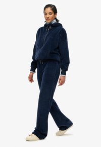 Granatowy dres z weluru z rozpinanym kapturem i spodniami na sznurki, o miękkiej fakturze i luźnym kroju. Połączony z jasnymi butami.