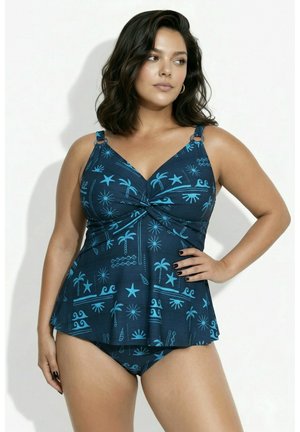 Woman wearing a dark blue two-piece swimsuit with light blue tropical and sea-themed patterns, posing with one hand on hip against a white background.