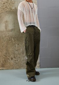 Person som bär en genomskinlig vit stickad tröja, olivgröna cargobyxor med lappar, svarta stövlar och ett pärlhalsband, stående inomhus.