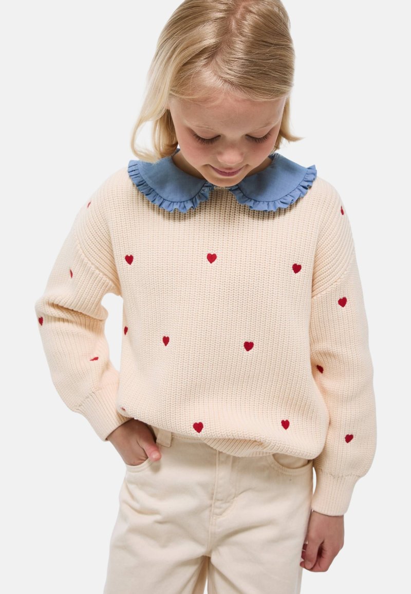 Pull en tricot crème avec des motifs de cœurs rouges et un col à volants bleu, associé à un pantalon beige clair. Tissu texturé avec une coupe décontractée.