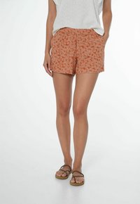 Dámské oranžové šortky s bílým květinovým vzorem, vyrobené z lehké tkaniny. Mají boční kapsy a volný střih. V kombinaci s hnědými sandály.