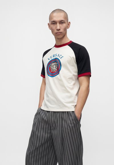Άνδρας μοντέλο με λευκό t-shirt της Versace με μαύρα μανίκια και κόκκινη περίγραμμα, συνδυασμένο με φαρδιά μαύρα και λευκά ριγέ παντελόνια.