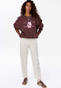 Žena s kudrnatými vlasy má na sobě hnědý svetr s motivem psa a židle, bílé kalhoty, růžové ponožky a hnědé tenisky, stojí se založenýma rukama za zády.